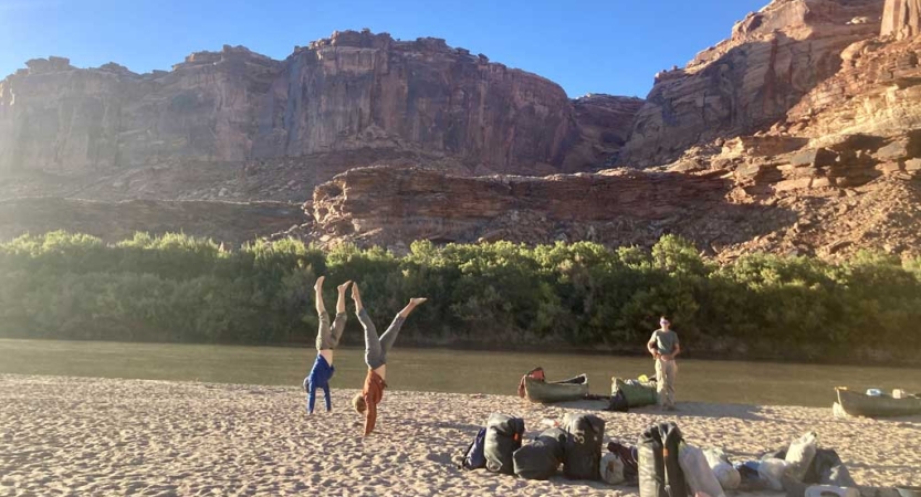 Two people perform a handstand on the sandy bank of a river. On the other side of the river are tall red canyon walls. 